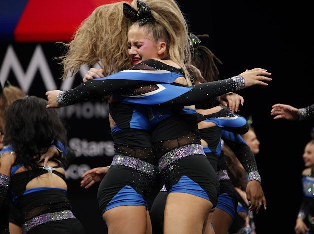 Cheerleaders in blue and black uniforms hugging, some crying, on a stage with