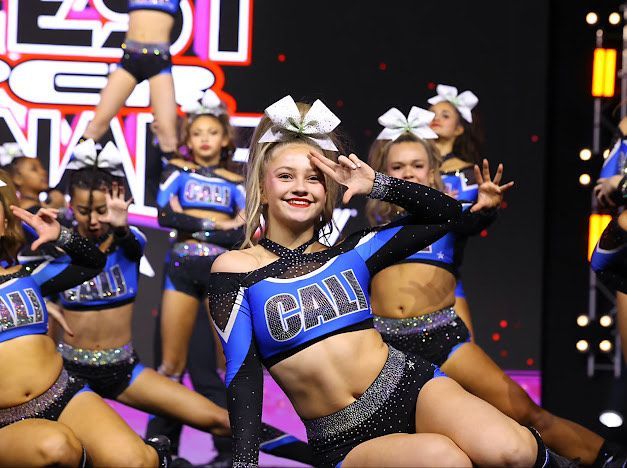 Cheerleaders in blue and black uniforms performing at a competition. Blonde girl smiles, making a hand gesture.