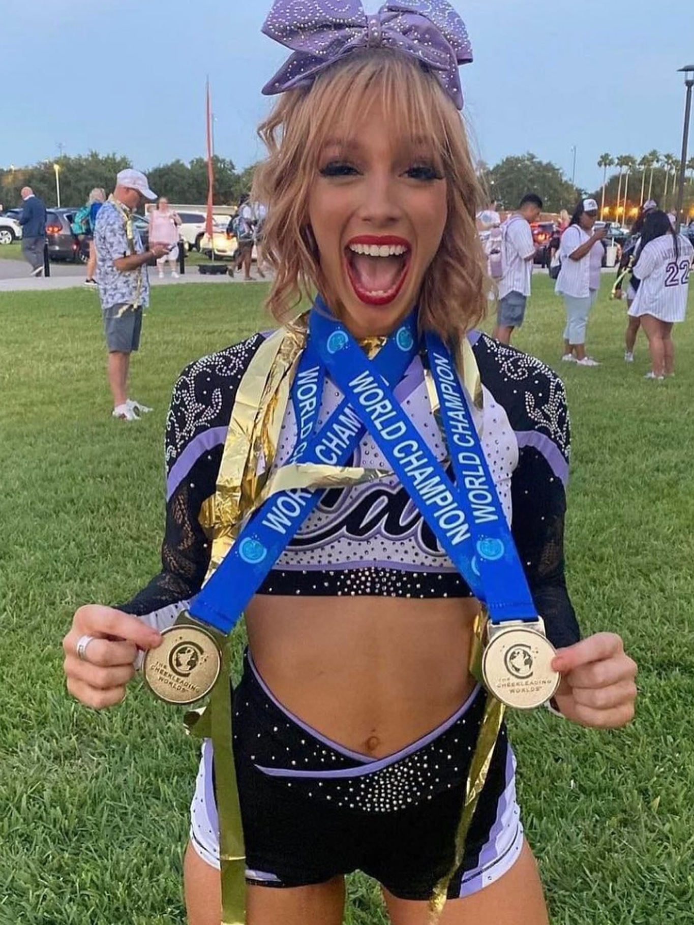 Cheerleader with gold medals, smiling excitedly. Purple, black, and gold uniform. Outdoor setting.