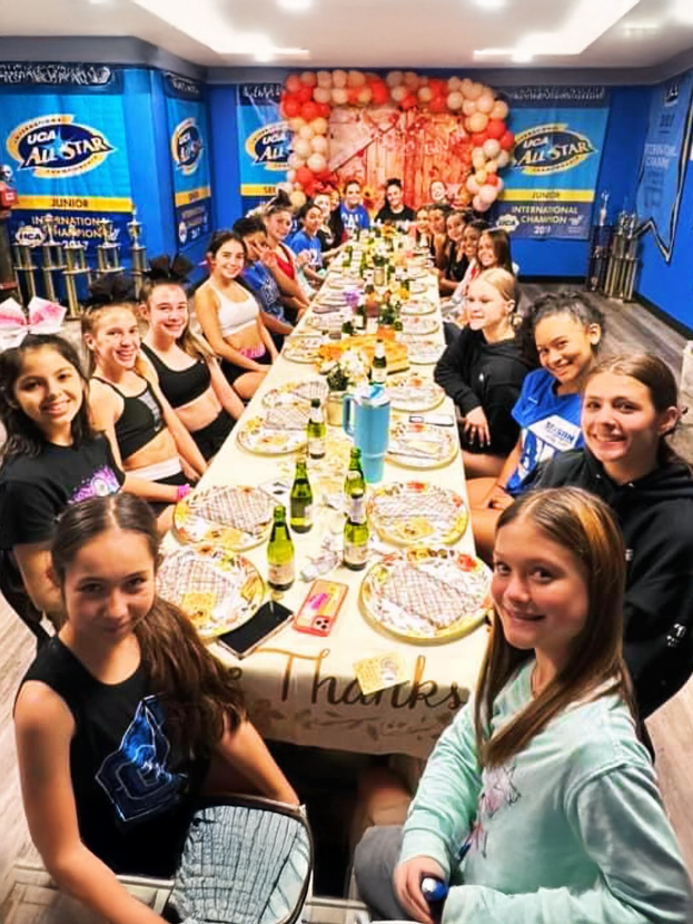 Group of cheerleaders posing in front of a banner. They are smiling, wearing athletic wear, and standing on a blue mat.