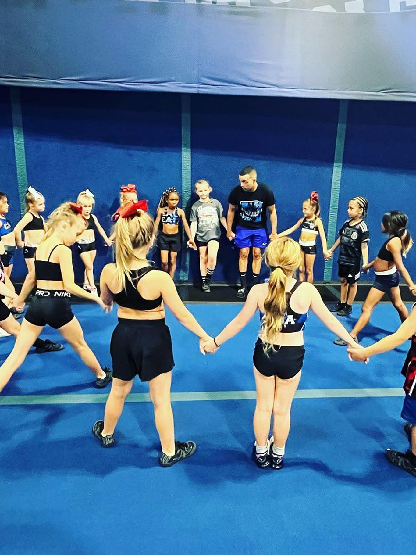A group of cheerleaders in black and blue uniforms holds hands in a circle around a male coach in a gym.