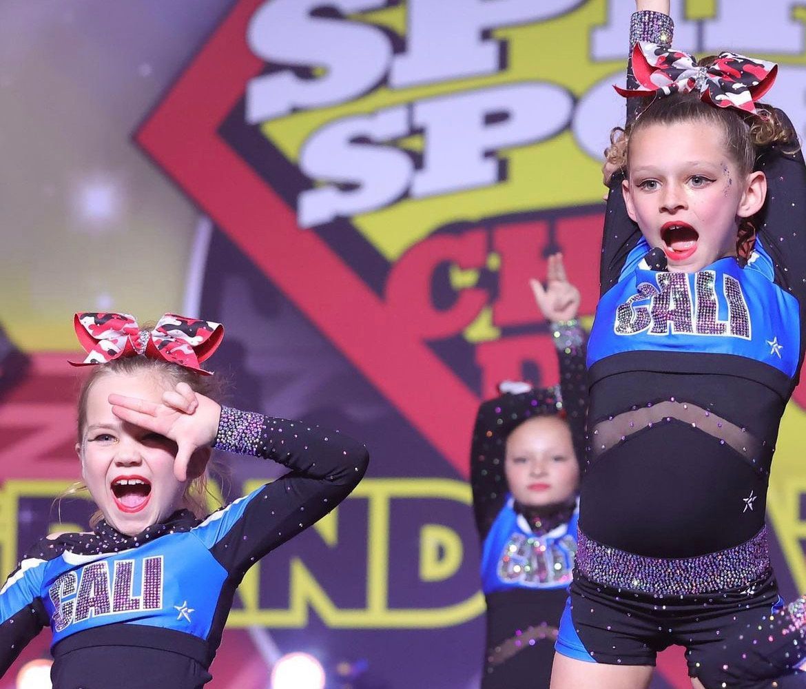 Cheerleaders in blue and black uniforms performing on a stage. Bright expressions, arm motions, and bows in hair.