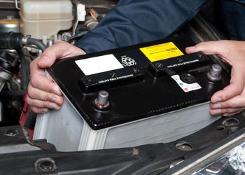 A person is holding a car battery under the hood of a car.