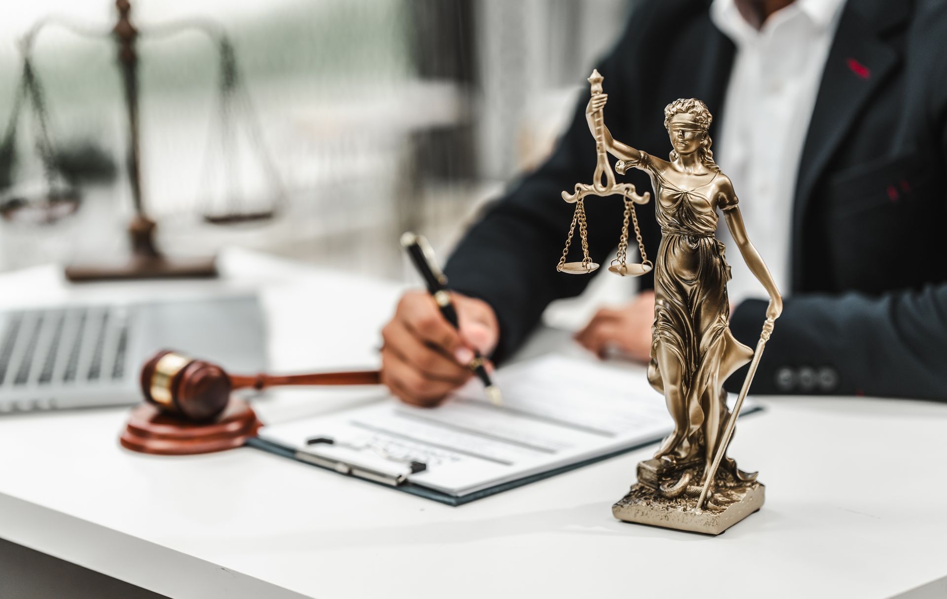 Lawyer signing documents at desk with Lady Justice statue, scales, and gavel.