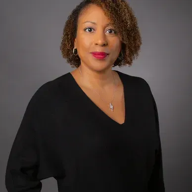 A woman wearing a black sweater and a necklace is standing in front of a gray background.