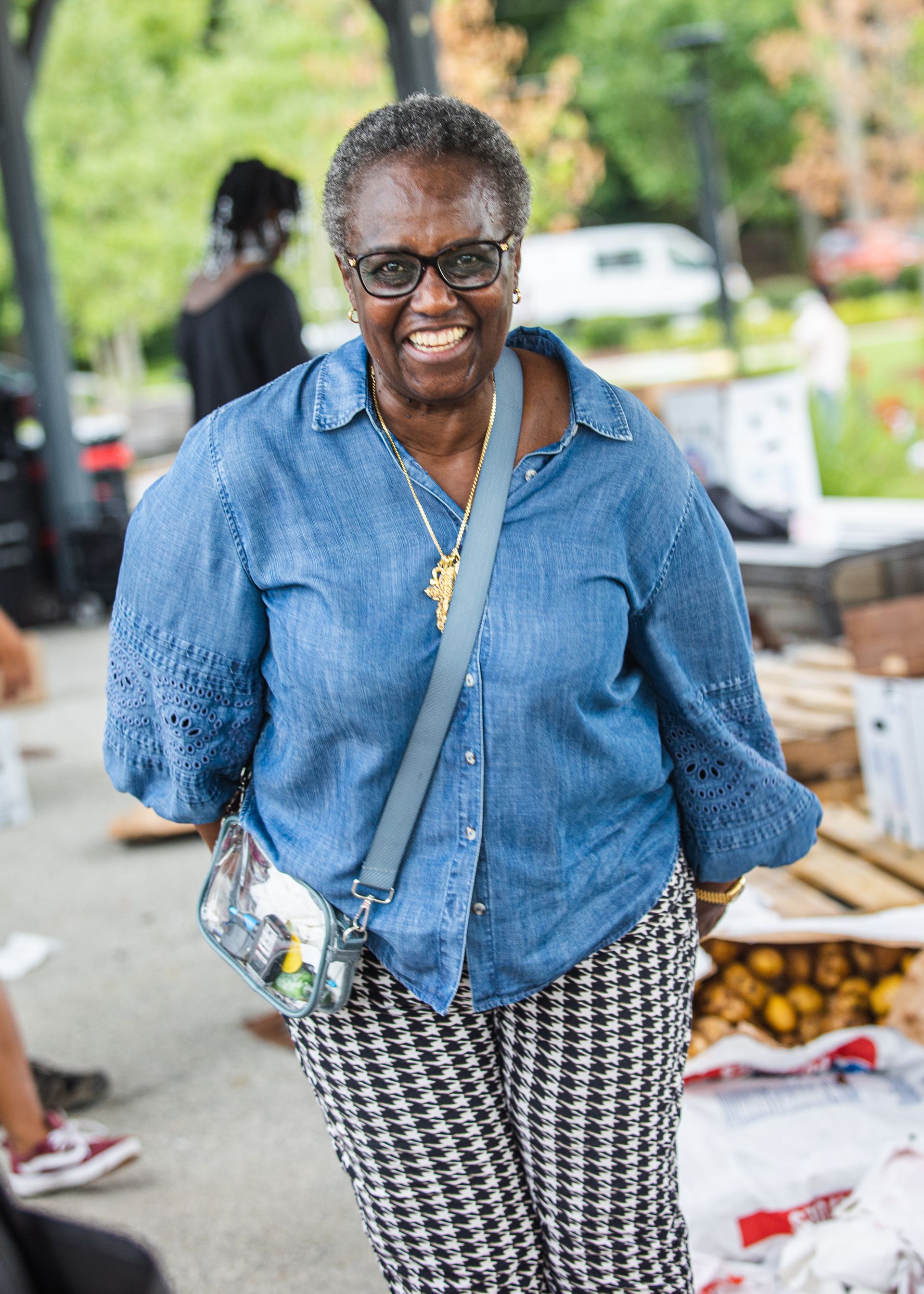 An elderly woman wearing glasses and a denim shirt is standing in a park.