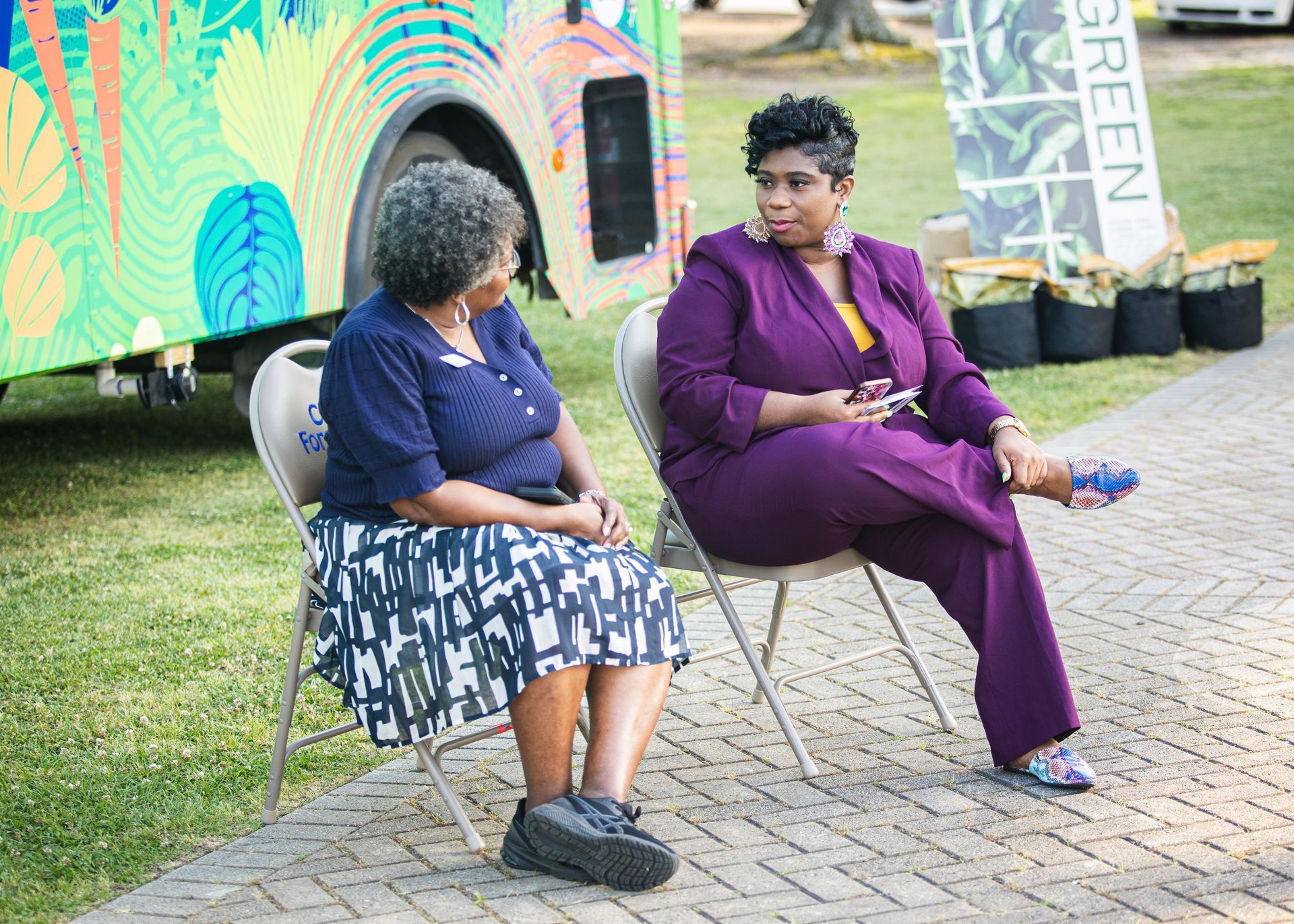 Two women are sitting in chairs in front of a colorful bus.