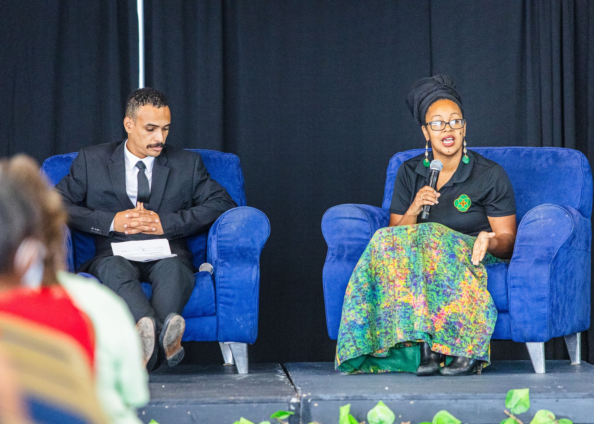 A man and a woman are sitting in blue chairs on a stage.