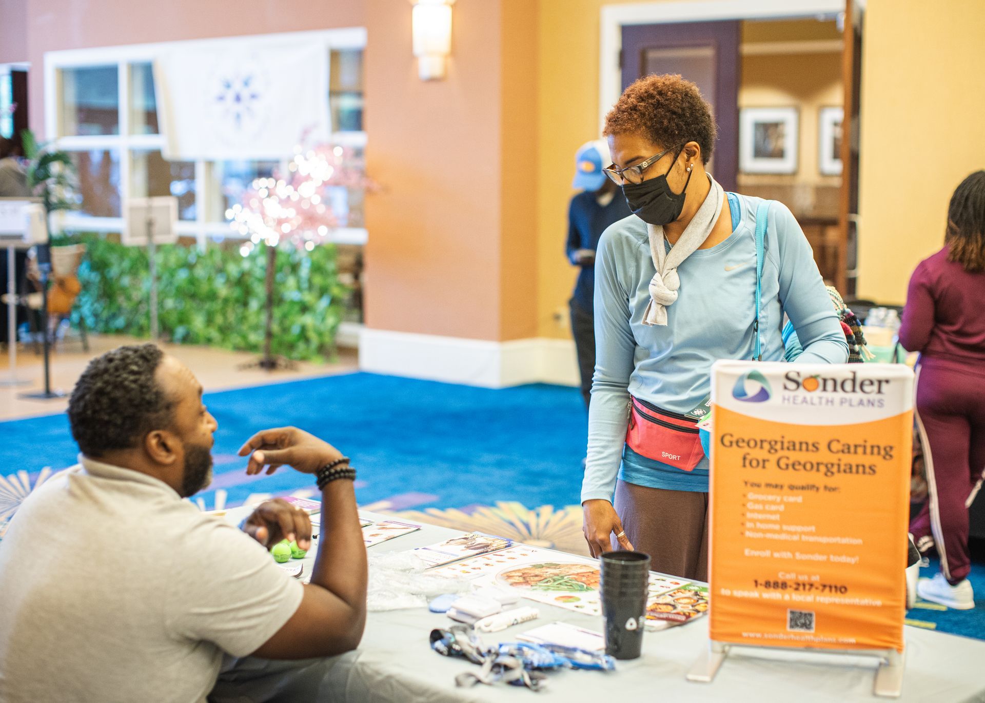 A woman wearing a mask is talking to a man at a table.