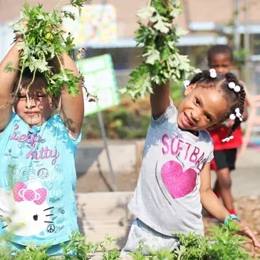 Two young girls are holding plants over their heads in a garden.