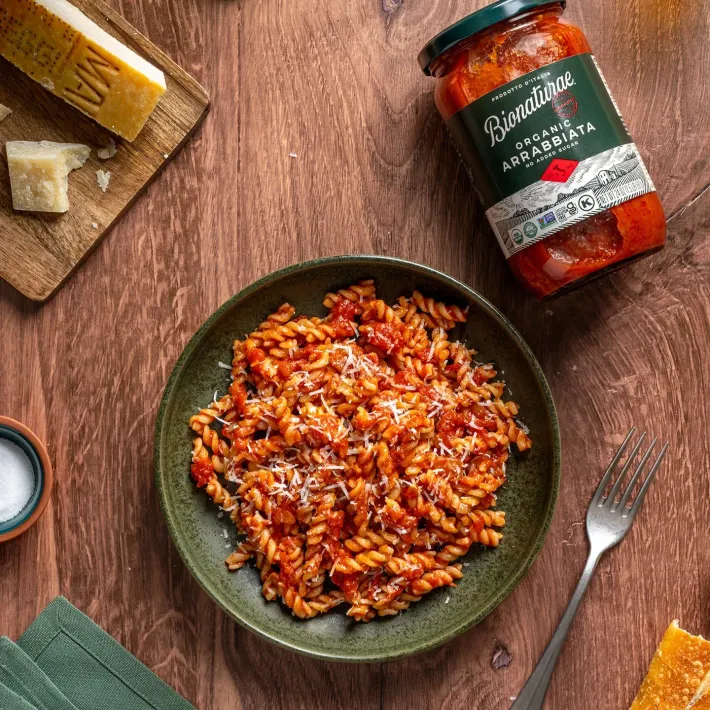 A bowl of fusilli pasta with arrabbiata sauce, next to a jar of Bionaturae sauce, cheese, and a fork on a wooden table.