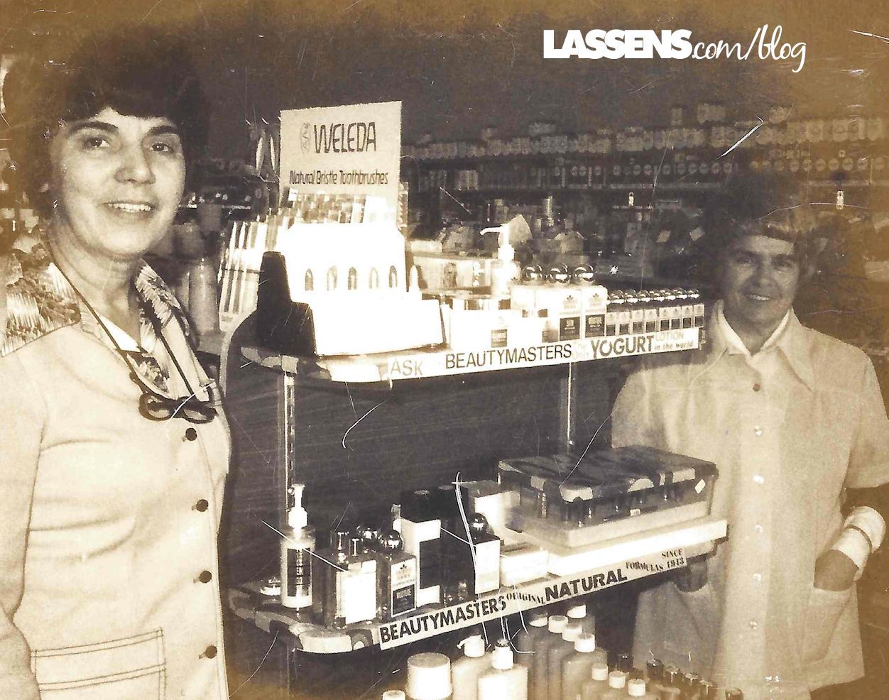 Two women stand inside a natural food store. One smiles, wearing a patterned shirt. Shelves stocked with products are visible.