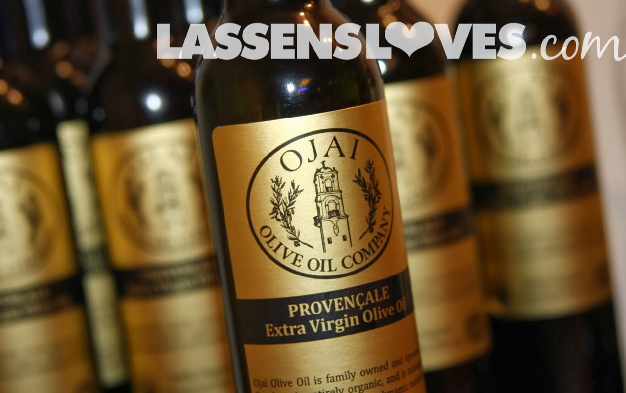Bottles of Ojai Olive Oil, with gold labels, on a shelf, lit from above.