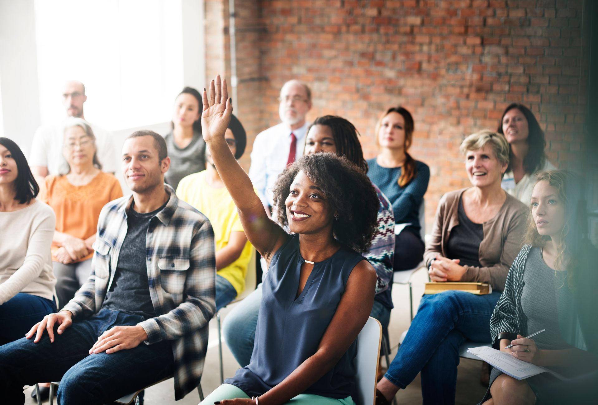 A woman with her hand raised in a workshop, surrounded by engaged participants in a room with a brick wall.