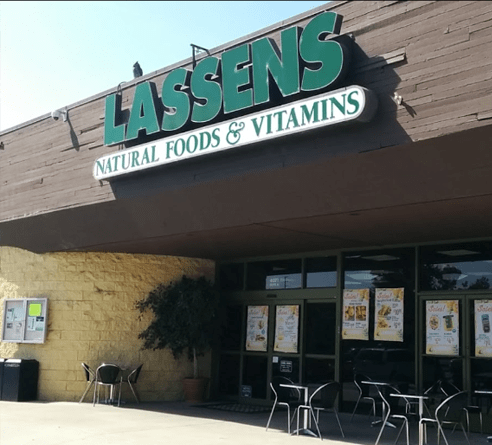 The storefront of Lassens Natural Foods & Vitamins with outdoor seating, a dark brown facade, and a green sign.