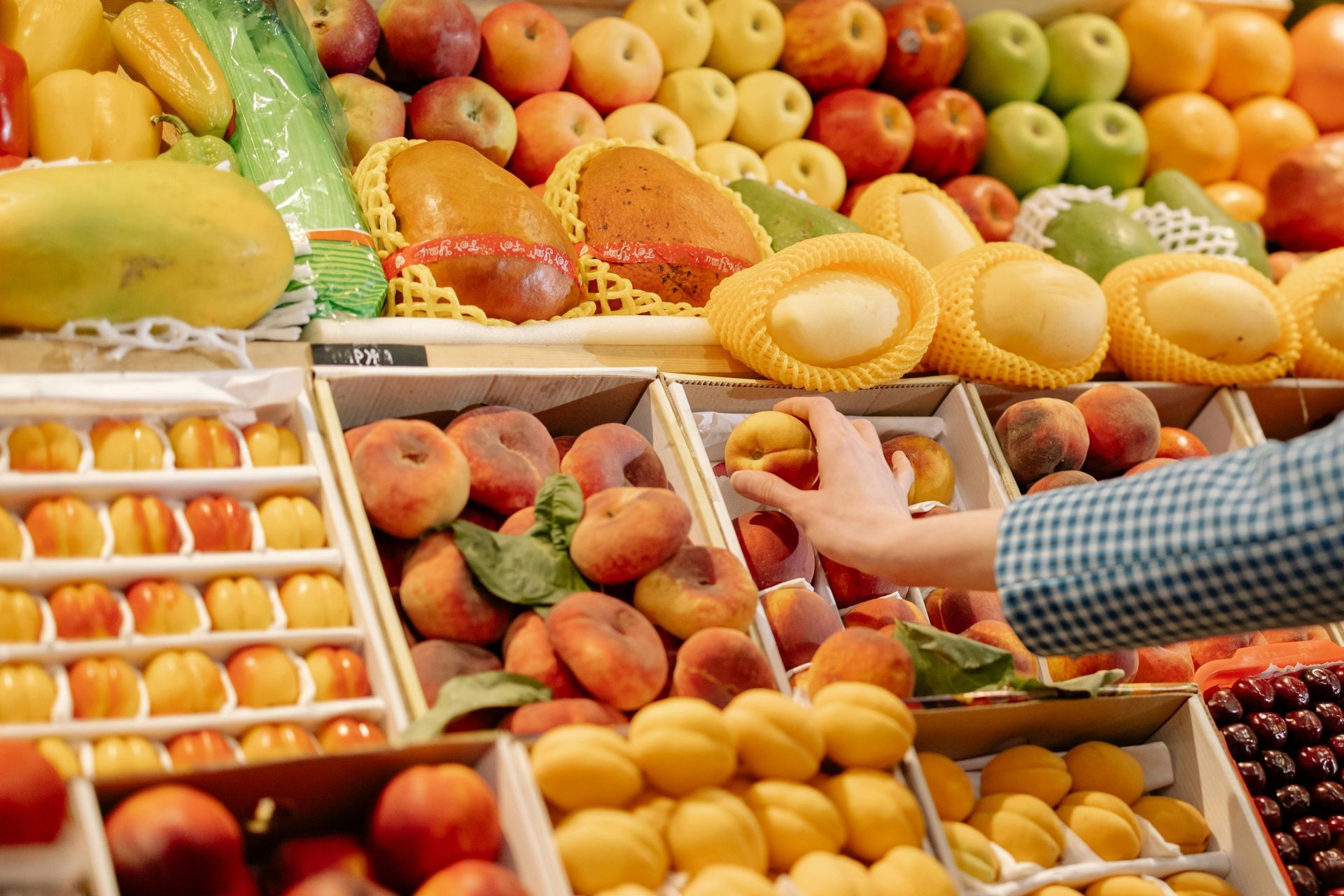 A hand selects a peach from a grocery display stocked with various fresh fruits including apples, mangoes, and cherries.