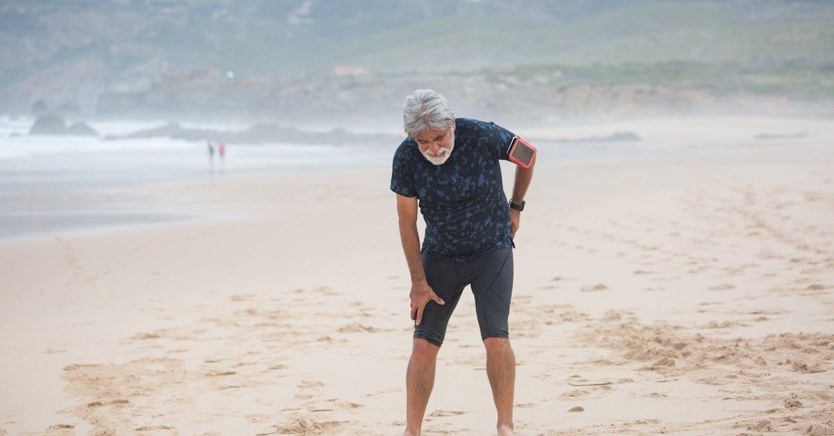 An older man is standing on a beach holding his knee in pain.