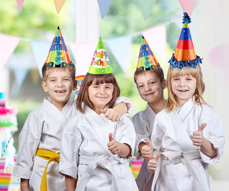 A group of children wearing karate uniforms and party hats are giving a thumbs up.