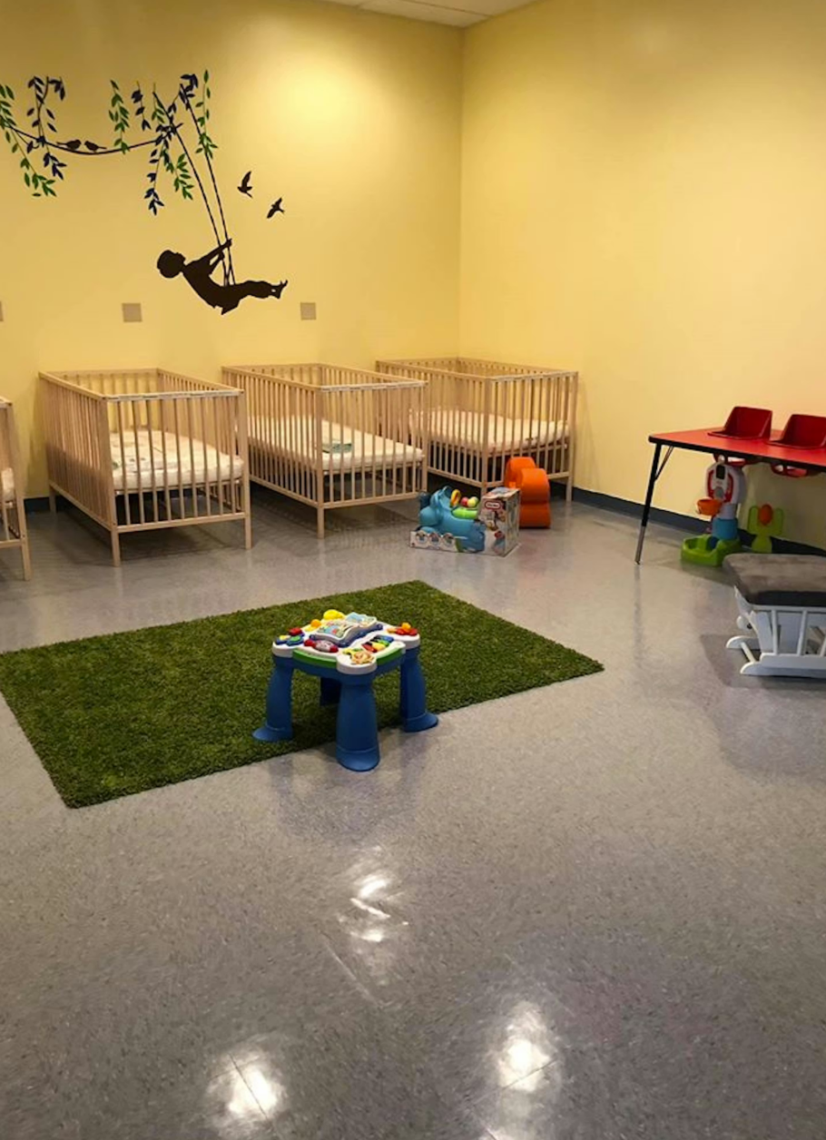 Nursery room with four cribs, a green rug, small table with toys, and wall mural of a child on a swing.