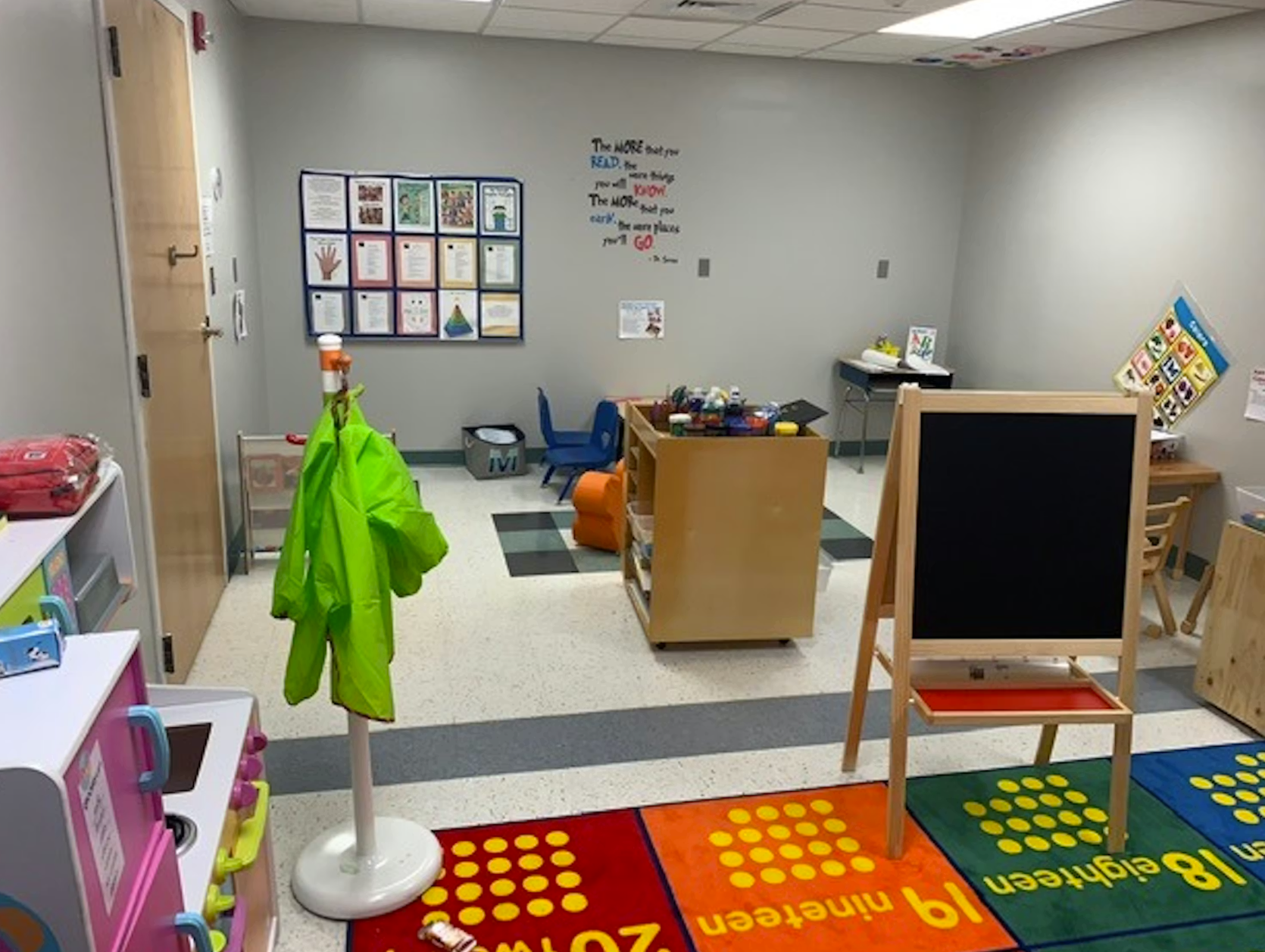 Classroom interior with a coat rack, easel, play kitchen, and bulletin board with children's artwork.