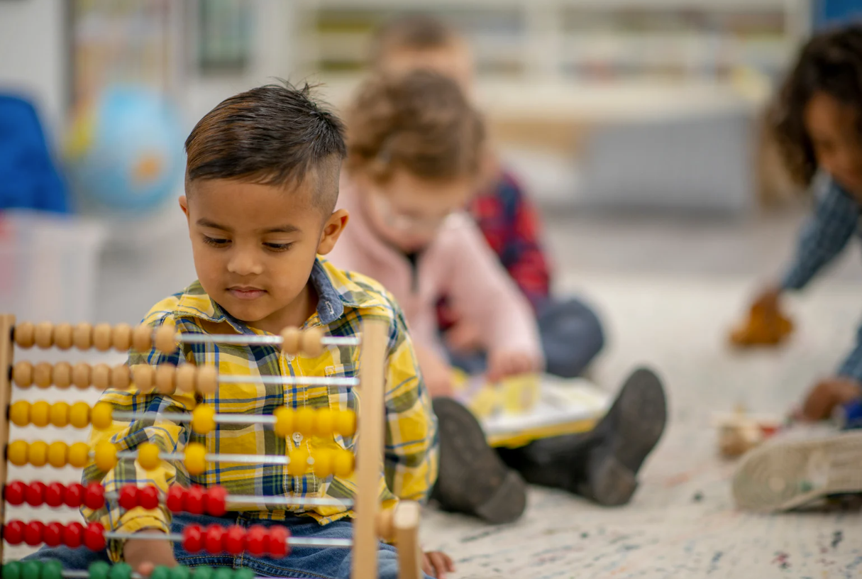 Boy using an abacus, other children in background. Classroom setting, focused expression, yellow plaid shirt.