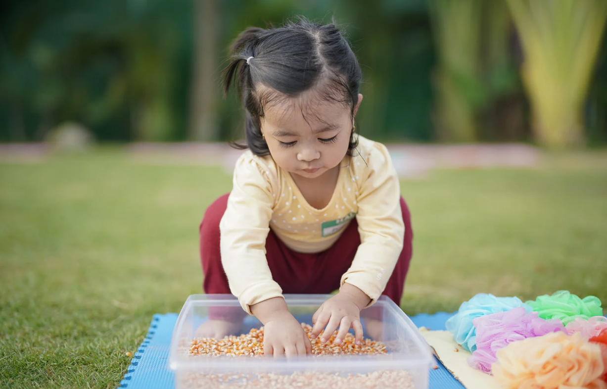 Child playing with grains in a clear container on grass, colorful objects nearby.