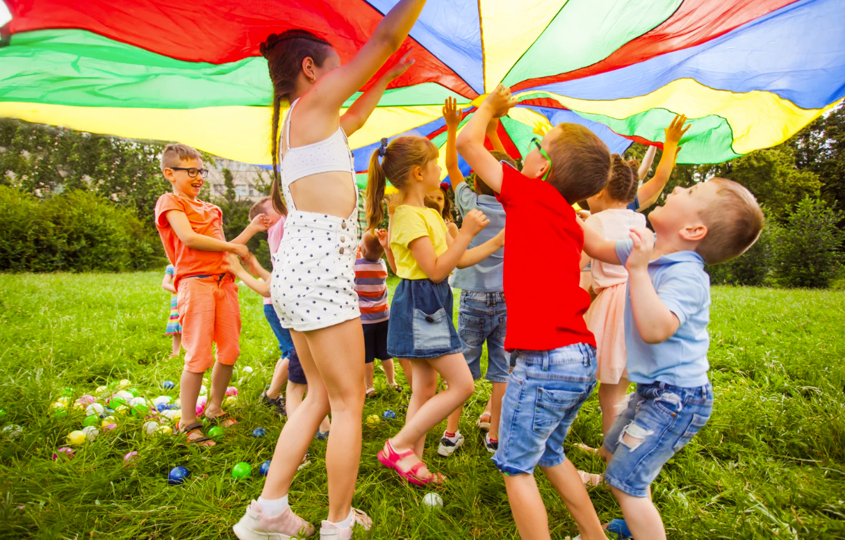Children playing with a colorful parachute on a grassy field, raising it up and down joyfully.