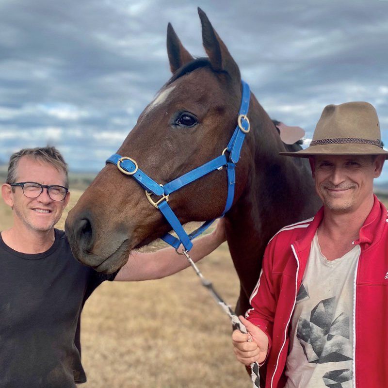 Two men standing next to a brown horse wearing a blue bridle