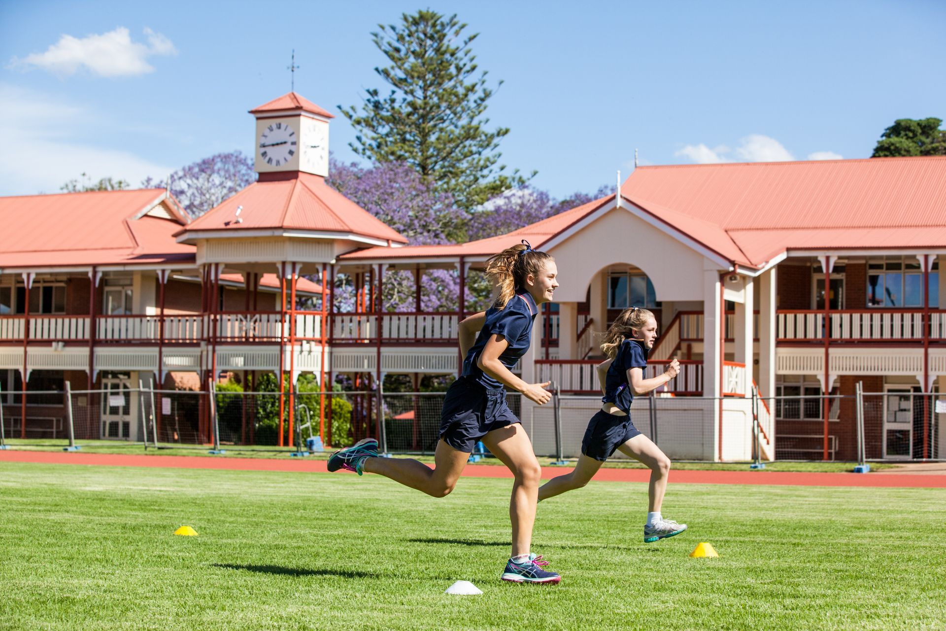 Two girls are running on a track in front of a building.