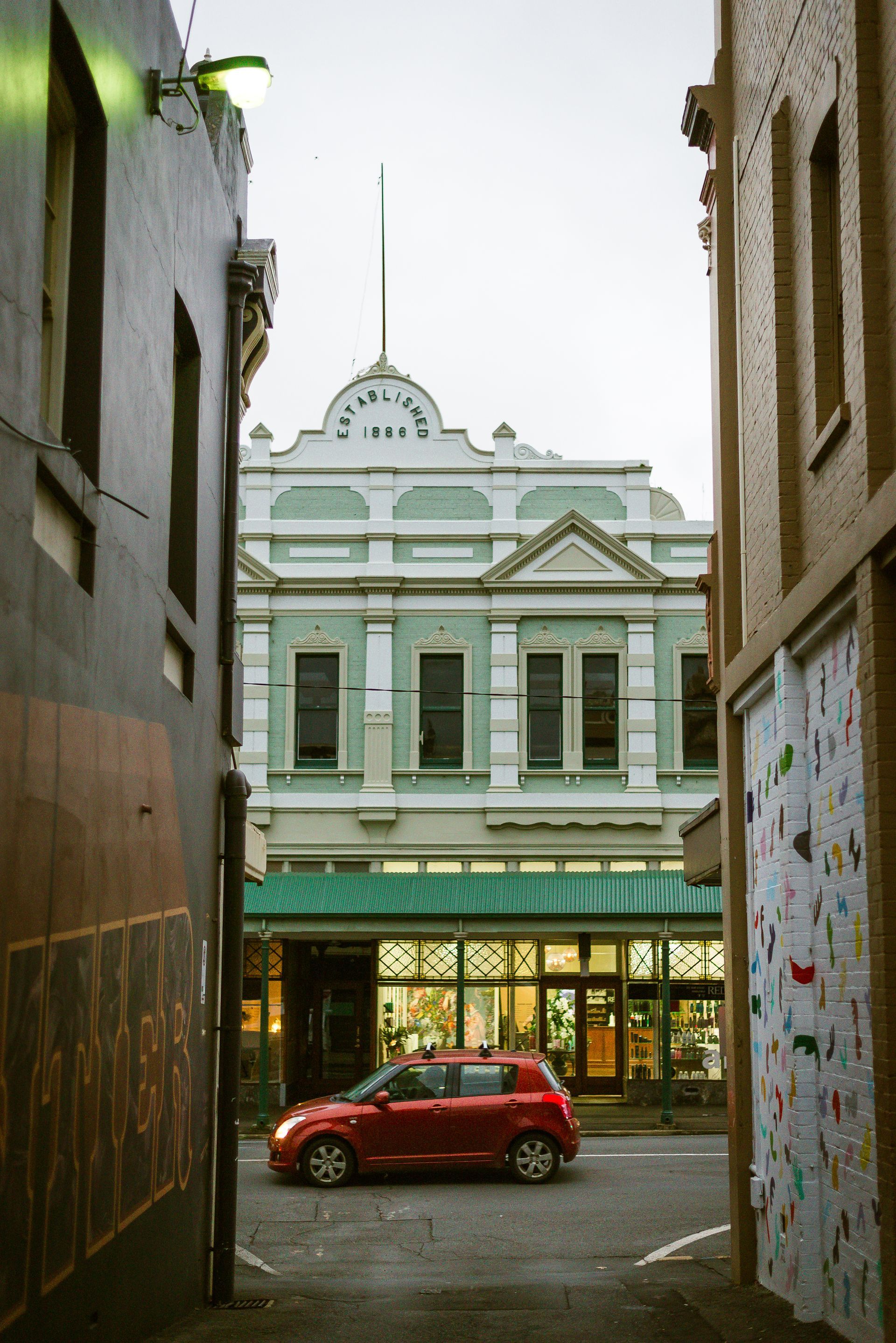 A red car is driving down a narrow street in front of a building