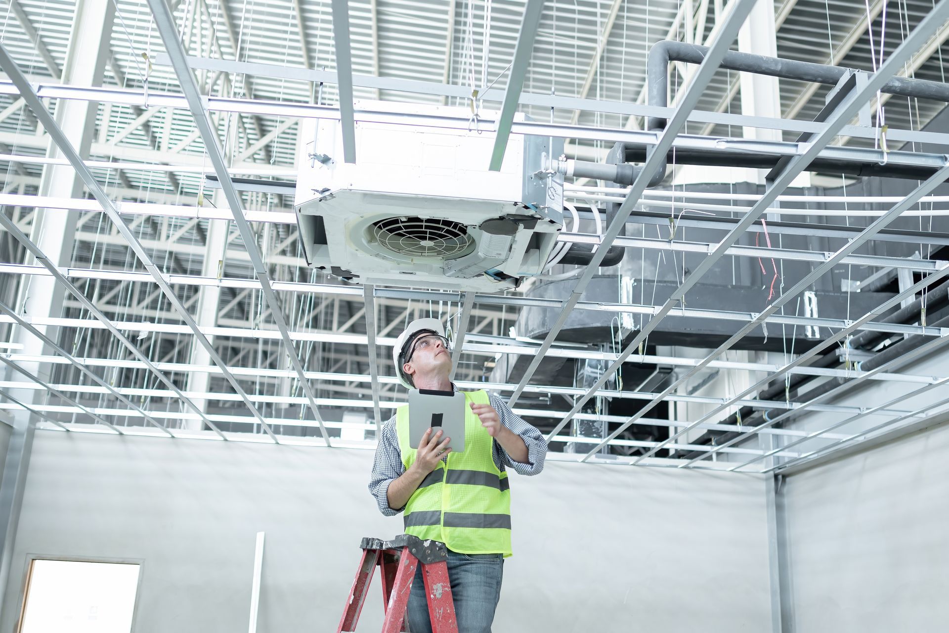 A construction worker is standing on a ladder looking up at a ceiling fan.