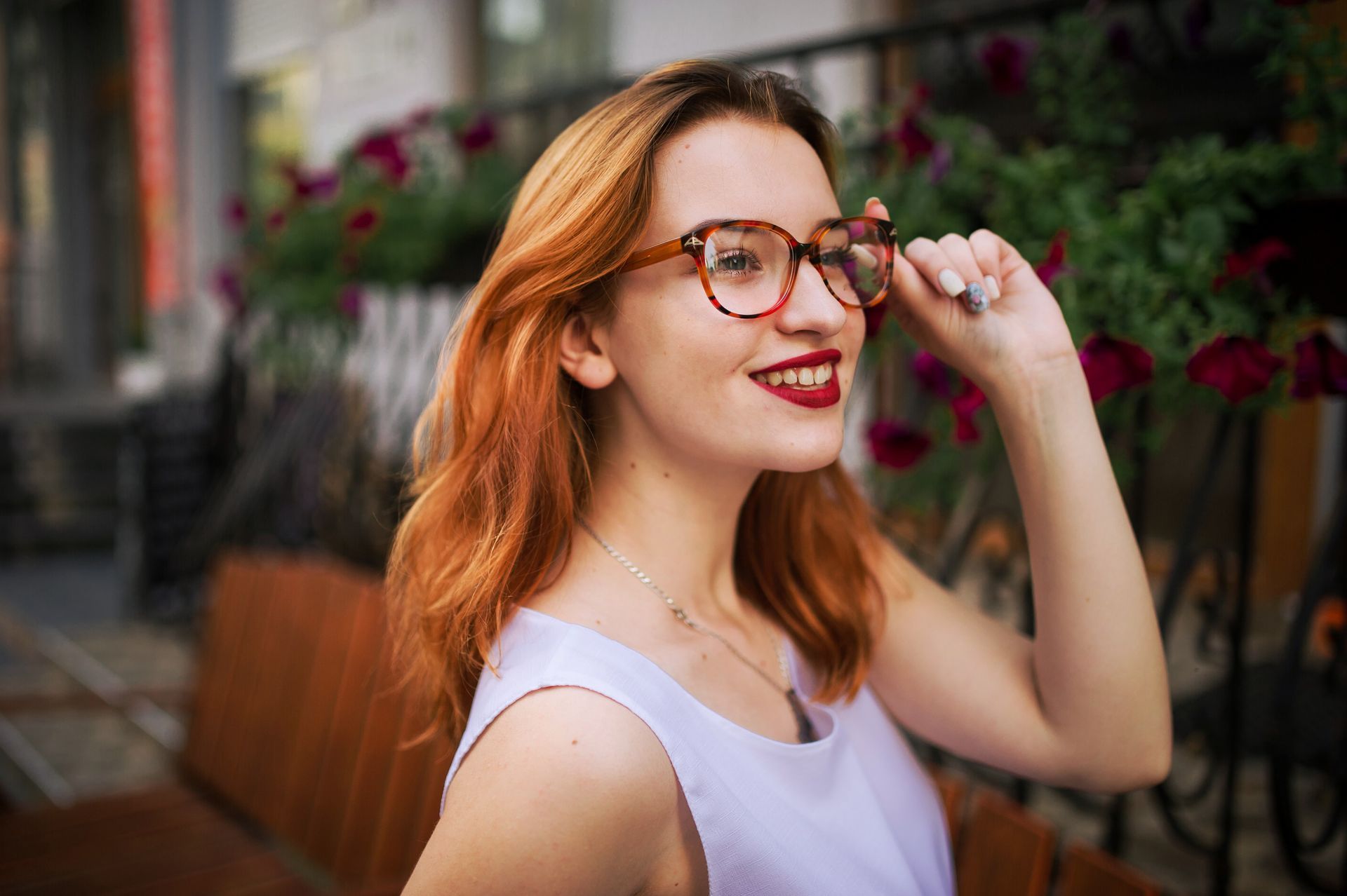 Woman with red hair and glasses smiles, touching her glasses. She wears a white top and stands outdoors near flowers and a bench.