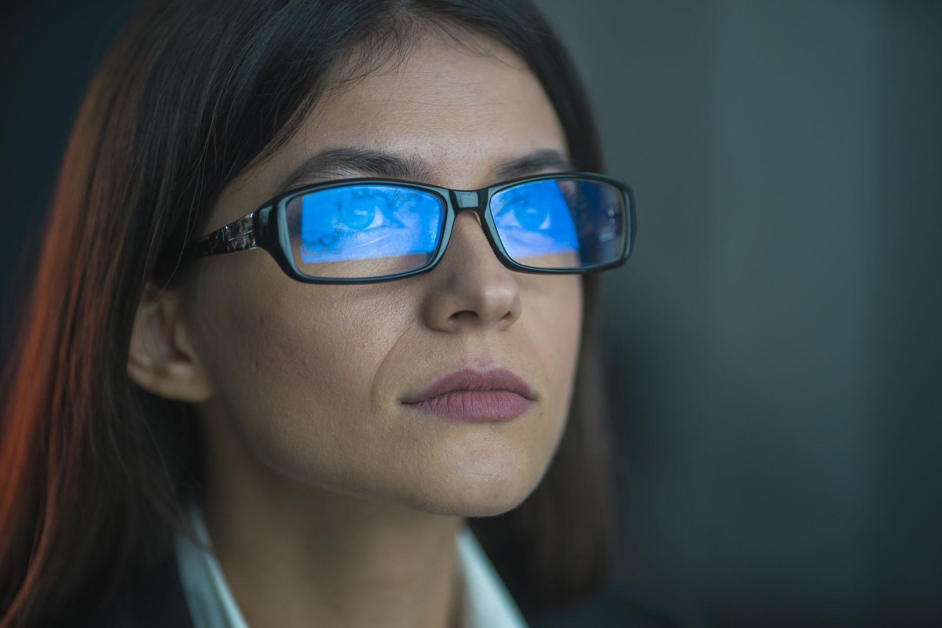 Woman wearing glasses with blue light reflection, looking intently, in a dark setting.