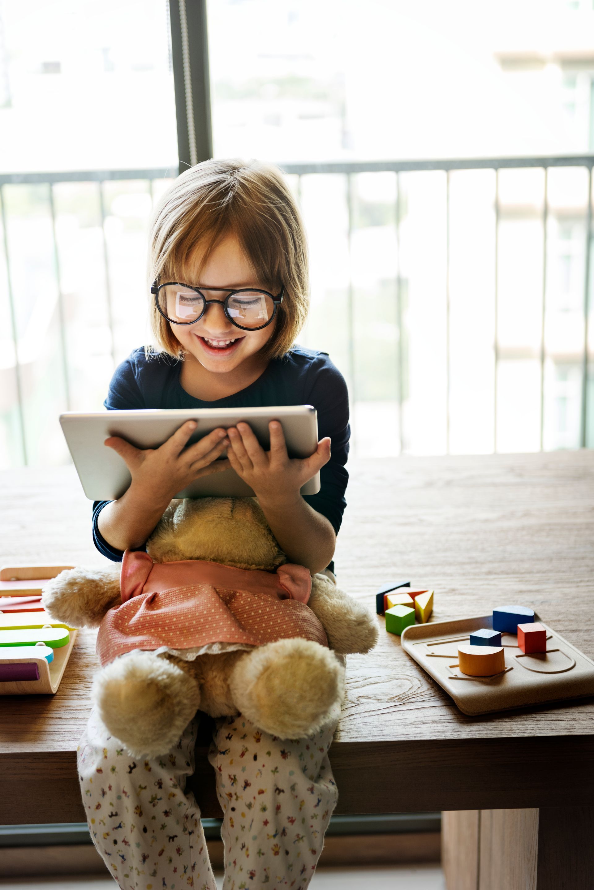 A little girl is sitting at a table with a teddy bear and a tablet.