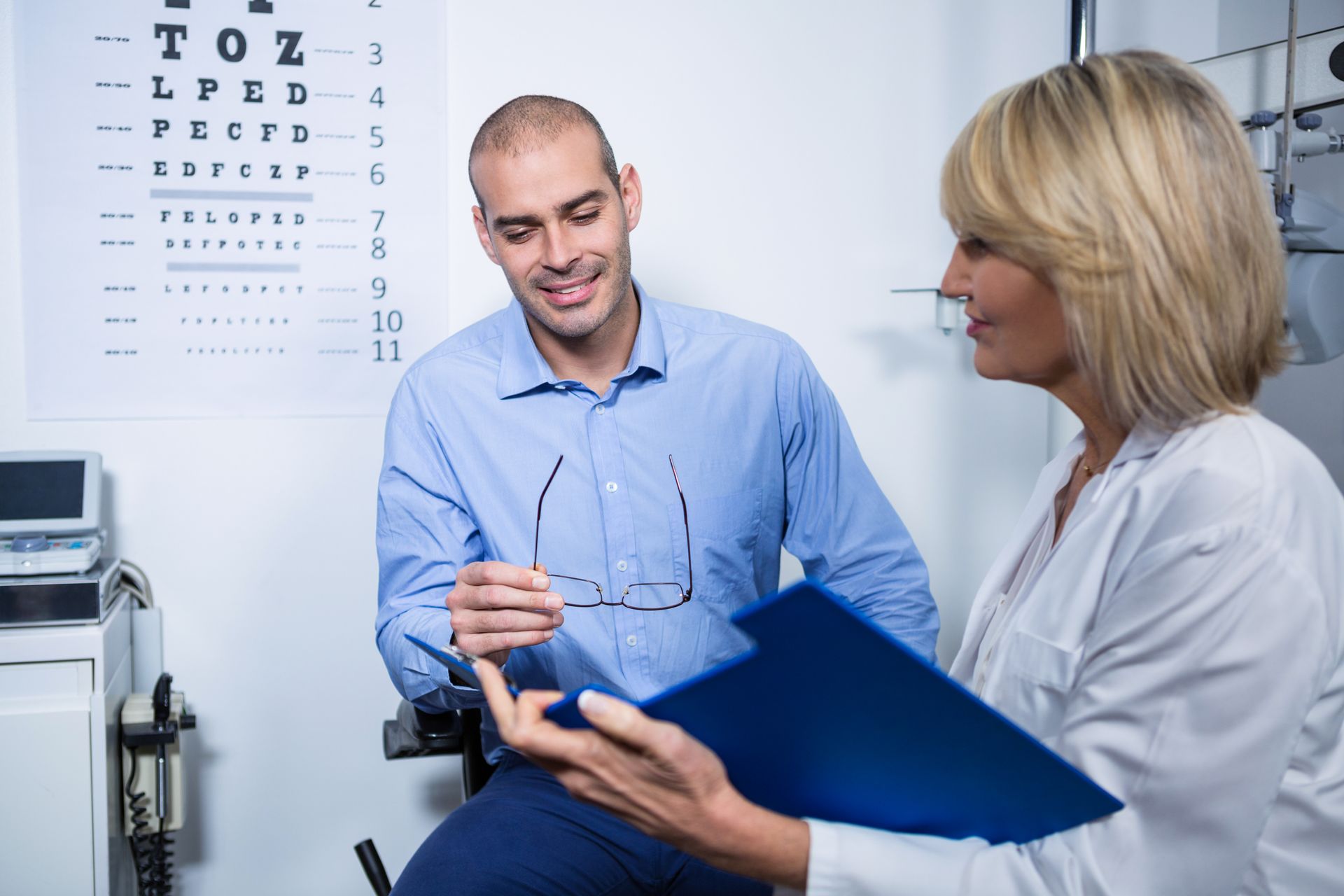 Man and eye doctor review results; eye chart visible. Man holds glasses, smiles; doctor holds clipboard.
