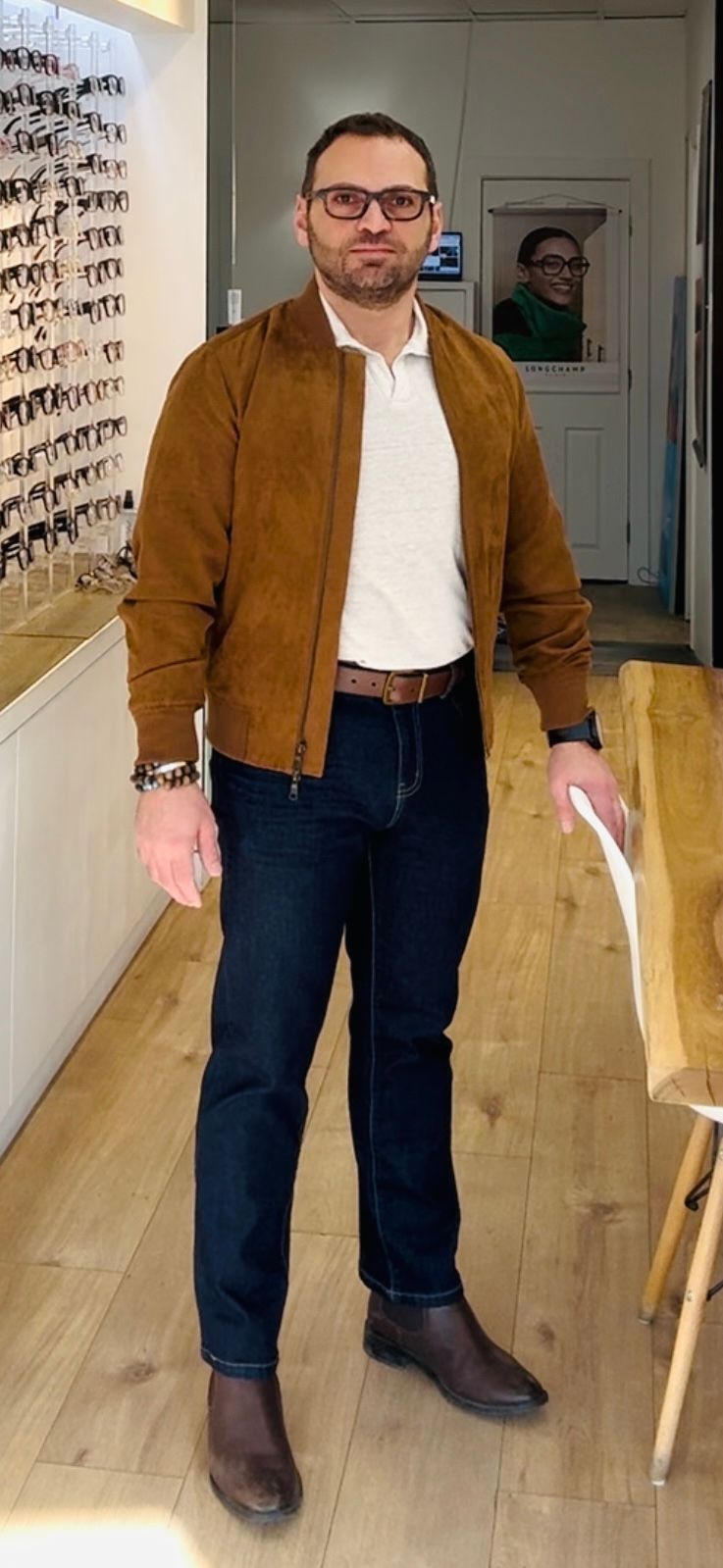 A man wearing glasses and a brown jacket is standing in an optical shop.