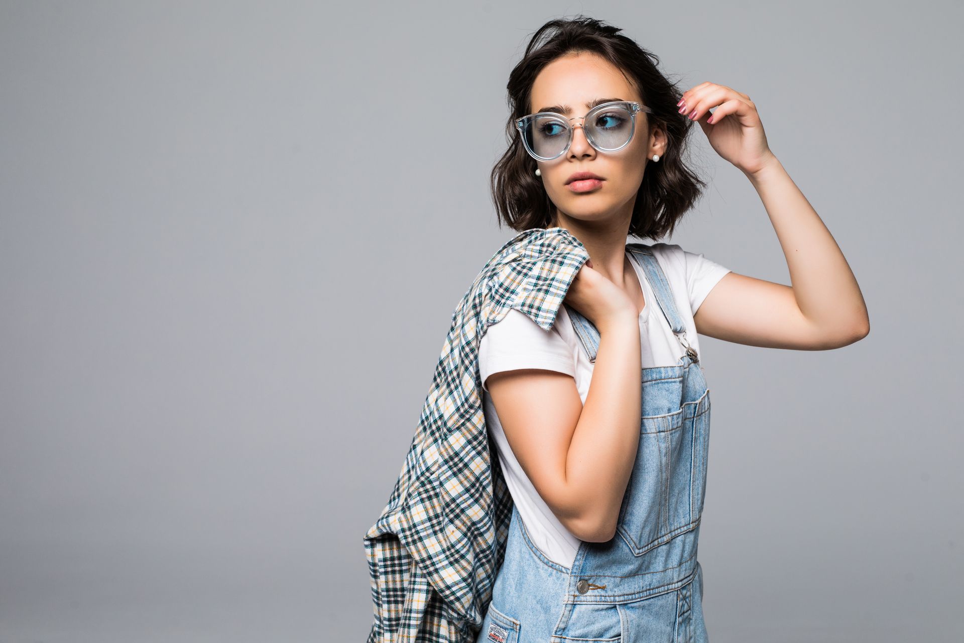 A woman wearing overalls and sunglasses is holding a plaid shirt over her shoulder.