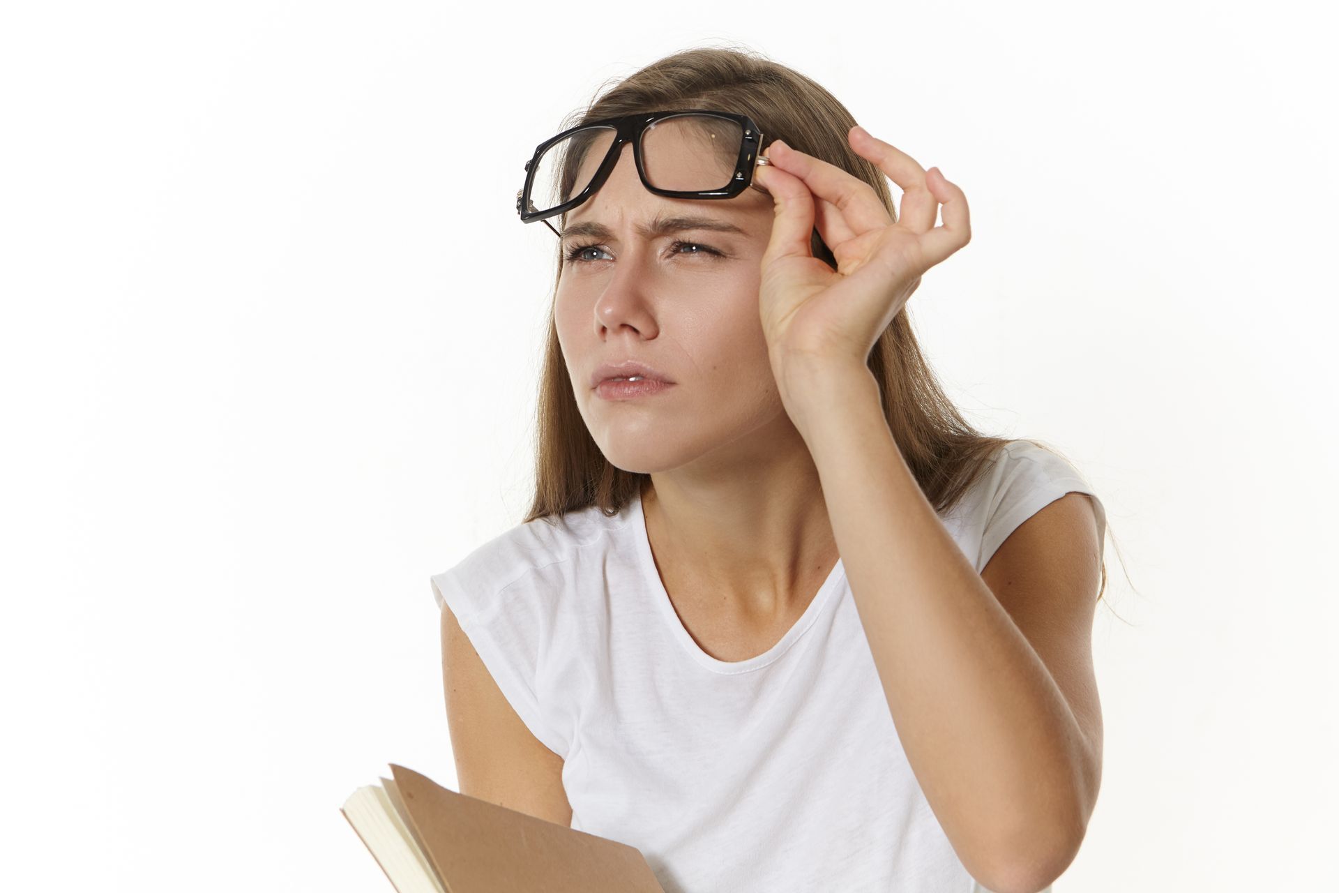 Woman in glasses squinting, holding book, white shirt, white background.
