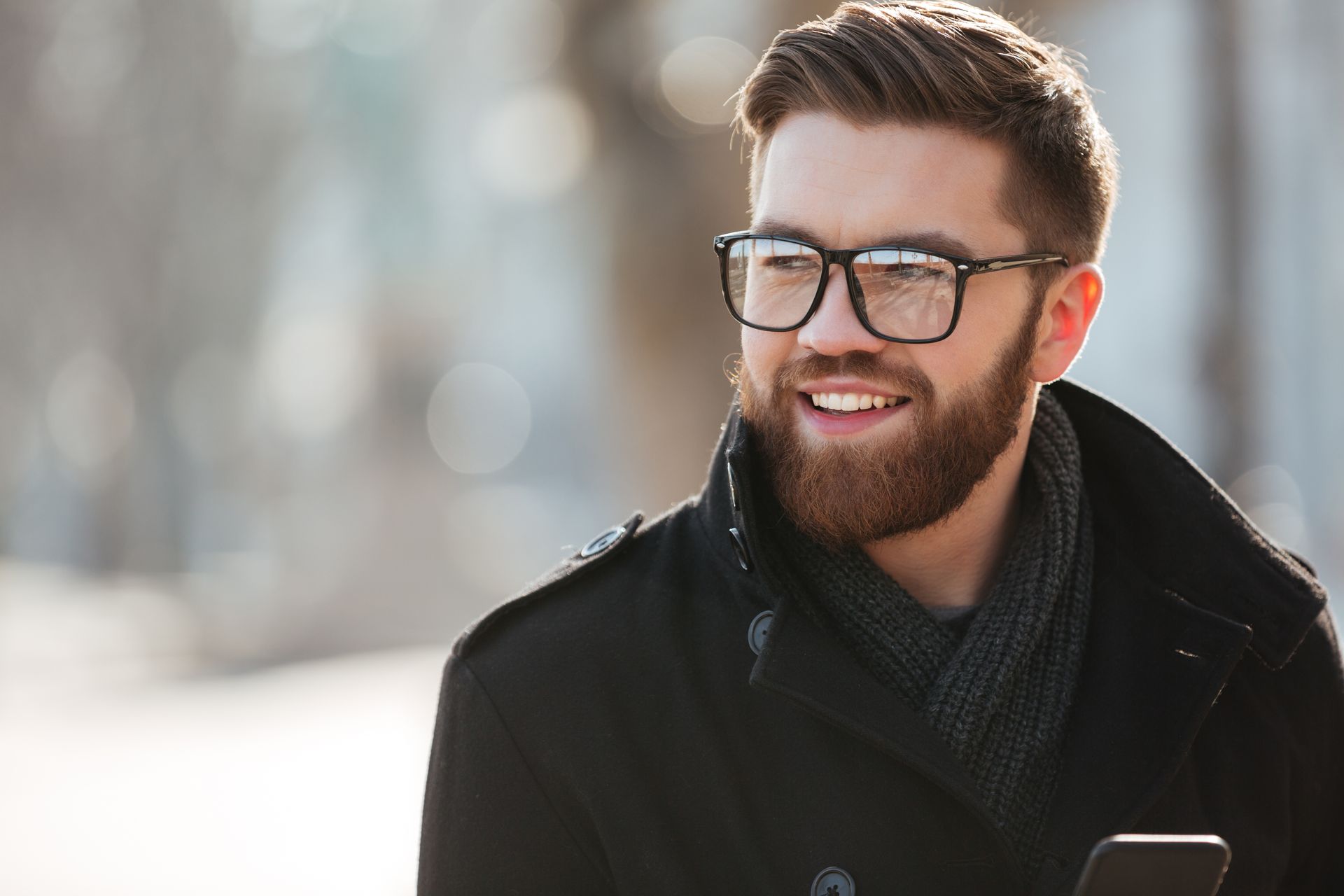 A man with a beard and glasses is smiling while holding a cell phone.