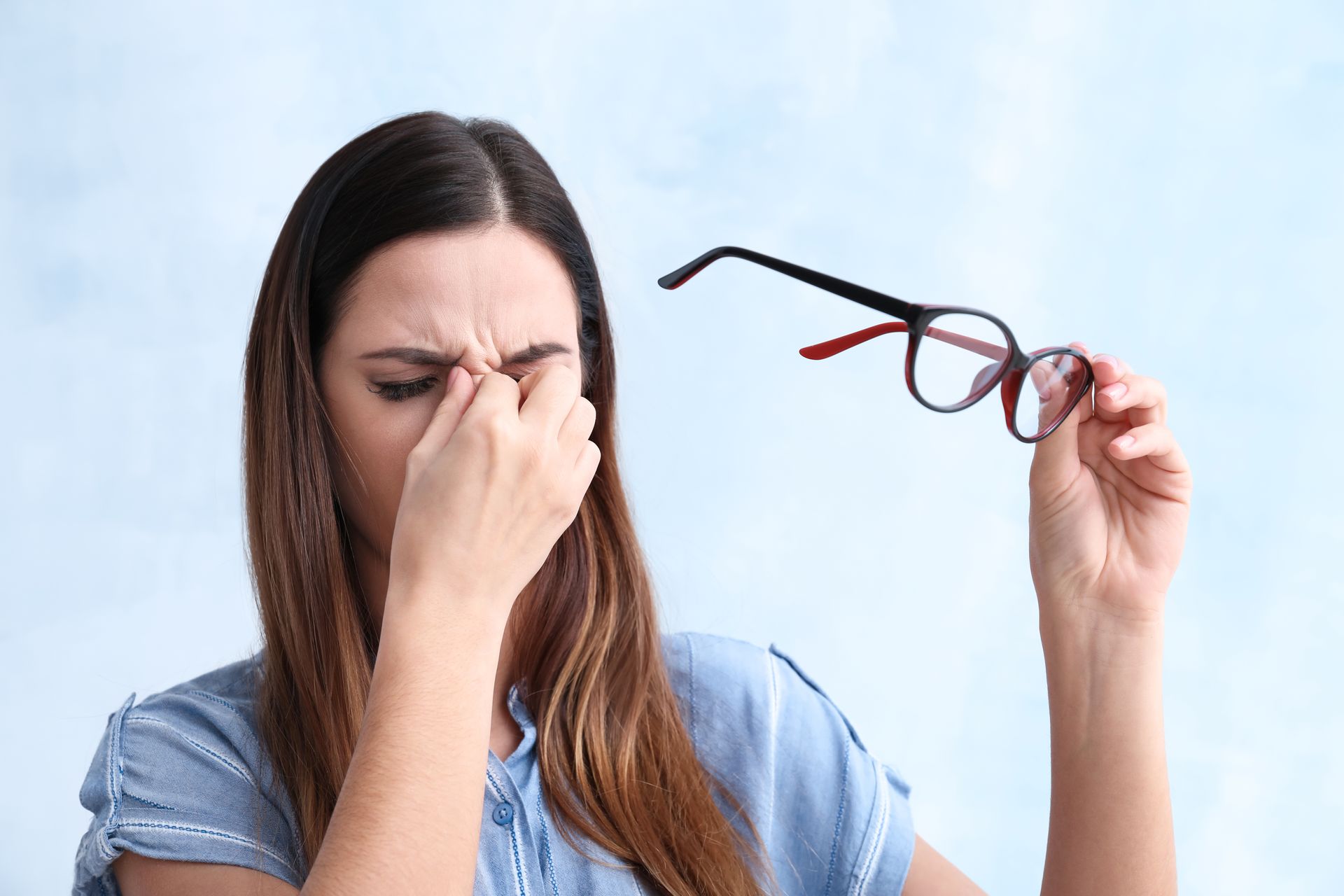 Woman holding glasses, pressing fingers to the bridge of her nose, looking strained. She has long brown hair, a light blue shirt, and a light blue background.
