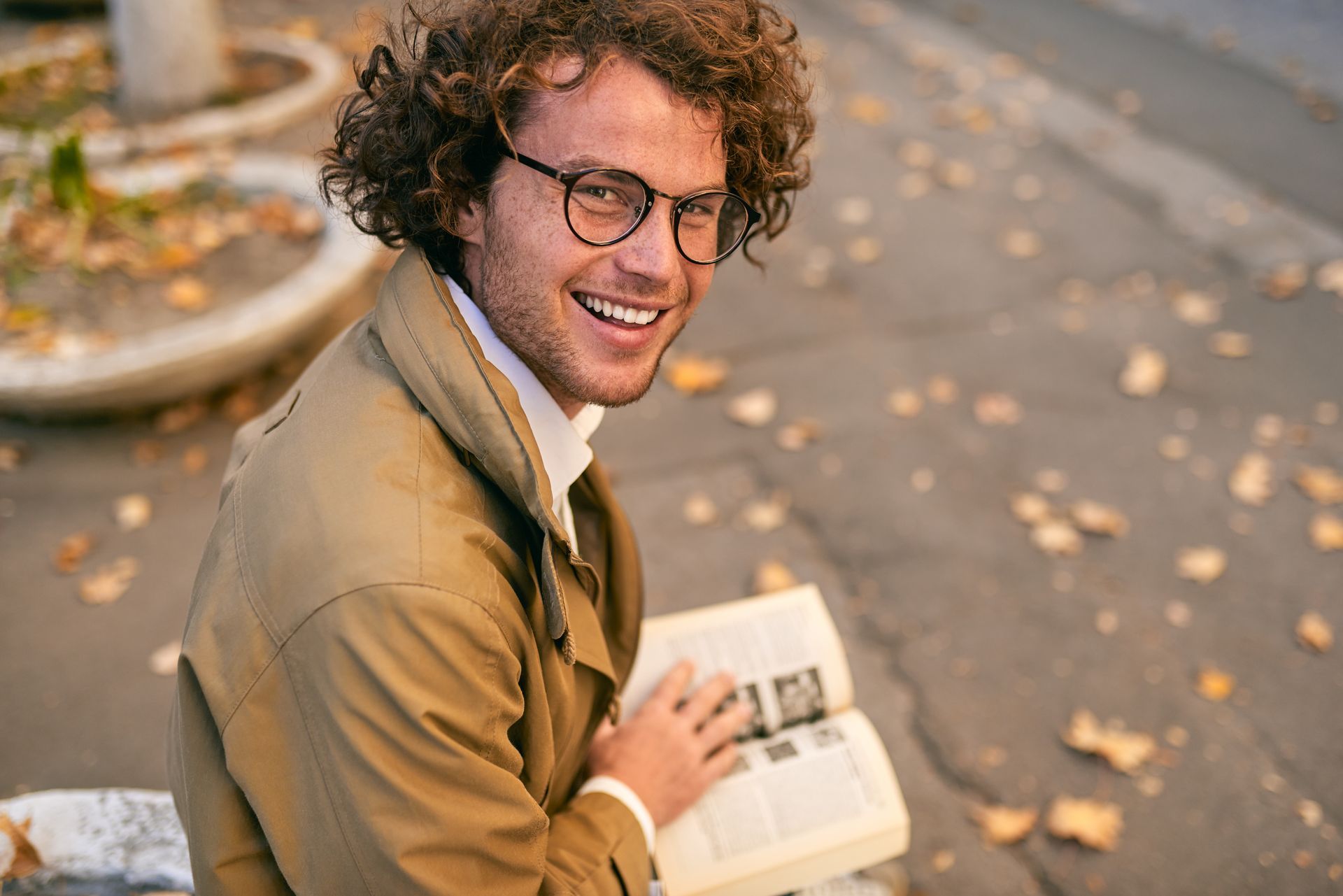 A smiling man with curly brown hair and glasses reads a book outdoors, wearing a tan coat.