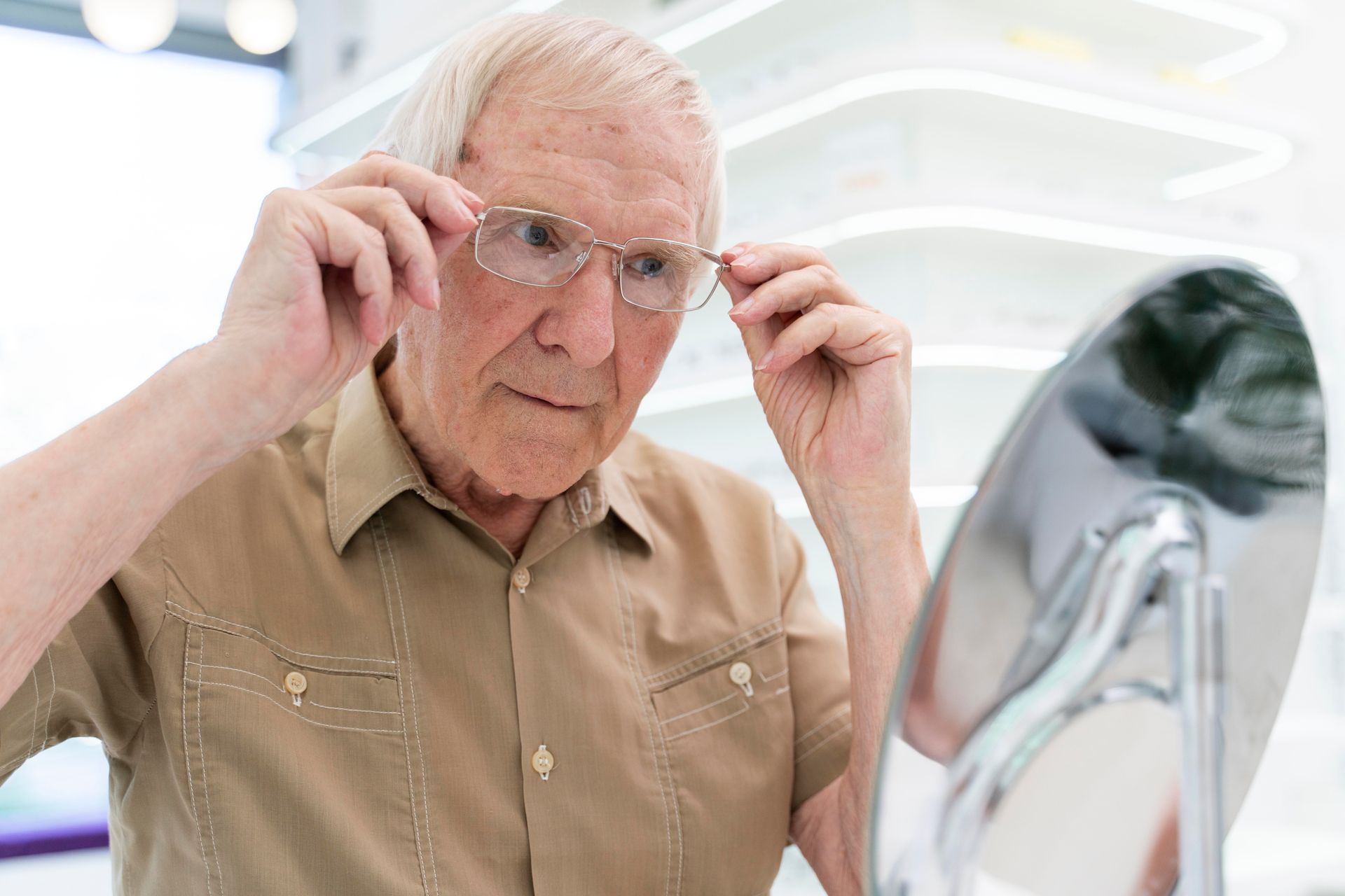 An older man in a tan shirt adjusts his glasses while looking in a mirror, possibly at an optical store.