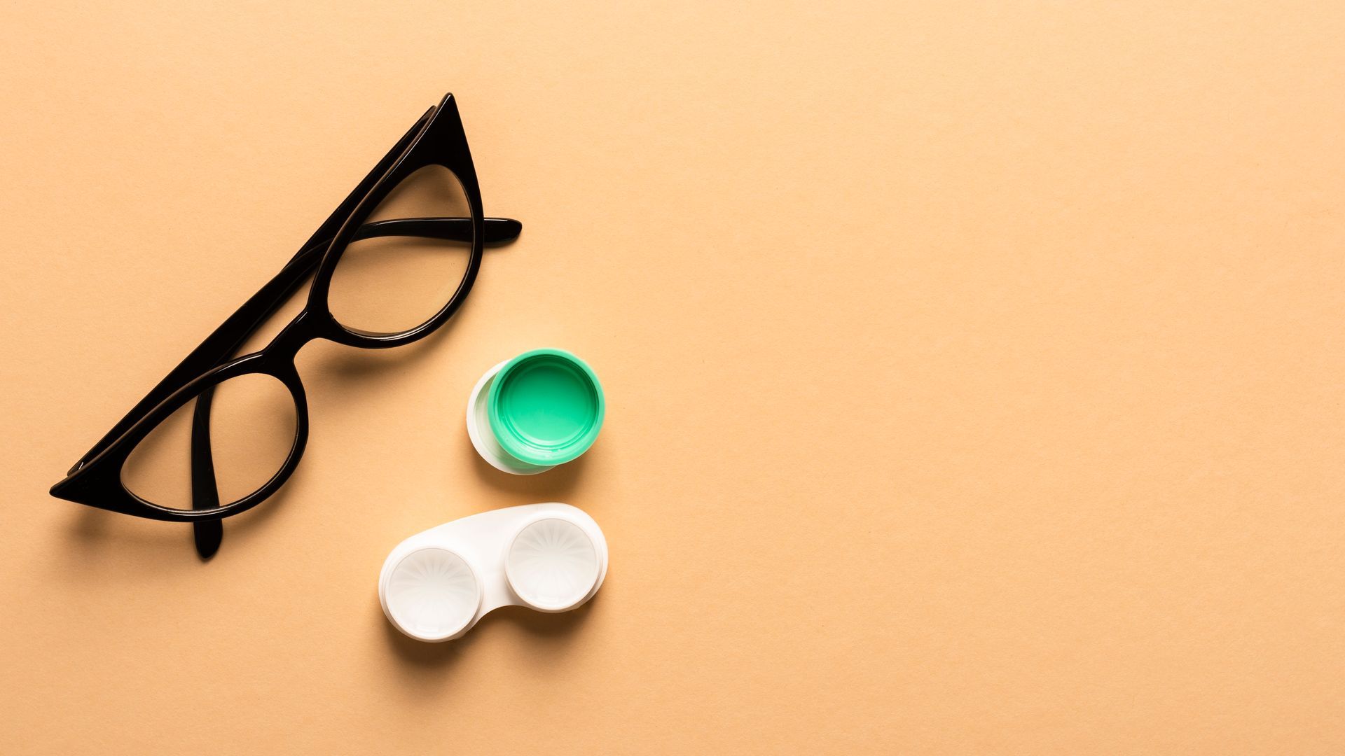 Black-framed eyeglasses and a green contact lens case are beside a white contact lens case on a light-orange surface.