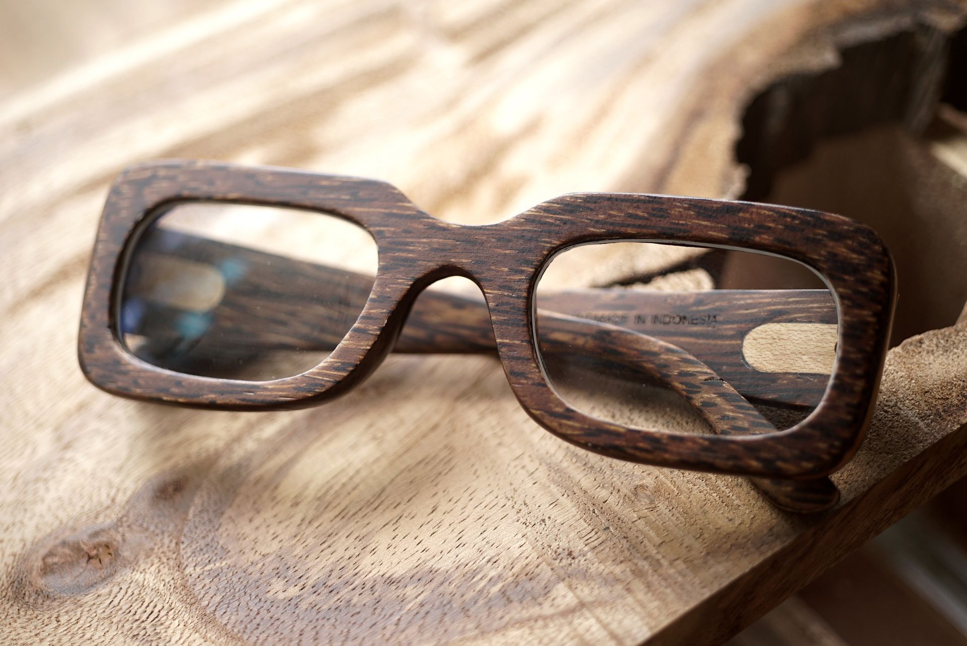 Wooden-framed eyeglasses resting on a piece of weathered wood. The glasses have rectangular lenses and a dark brown finish.