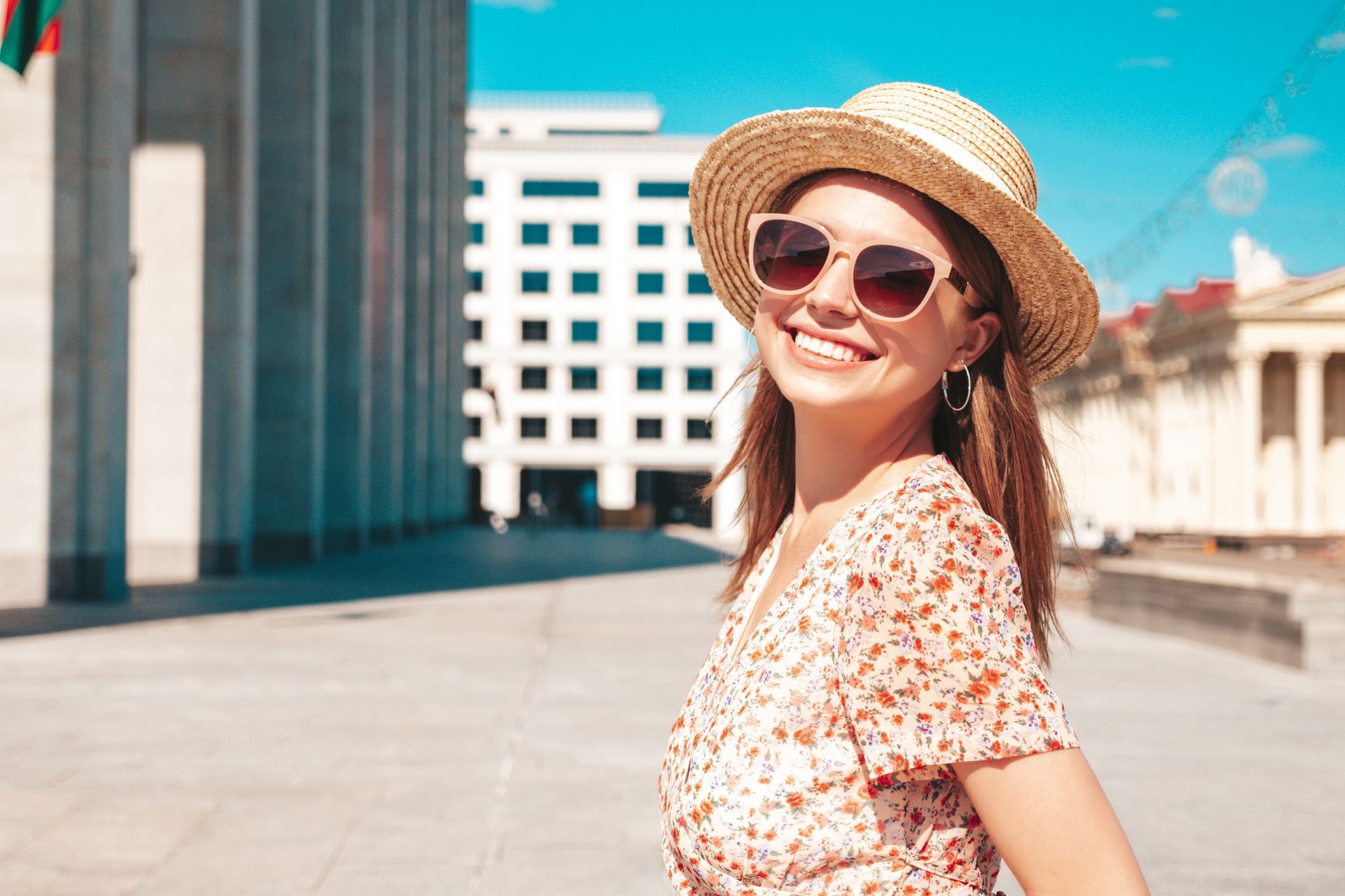 A woman wearing a straw hat and sunglasses is smiling in front of a building.