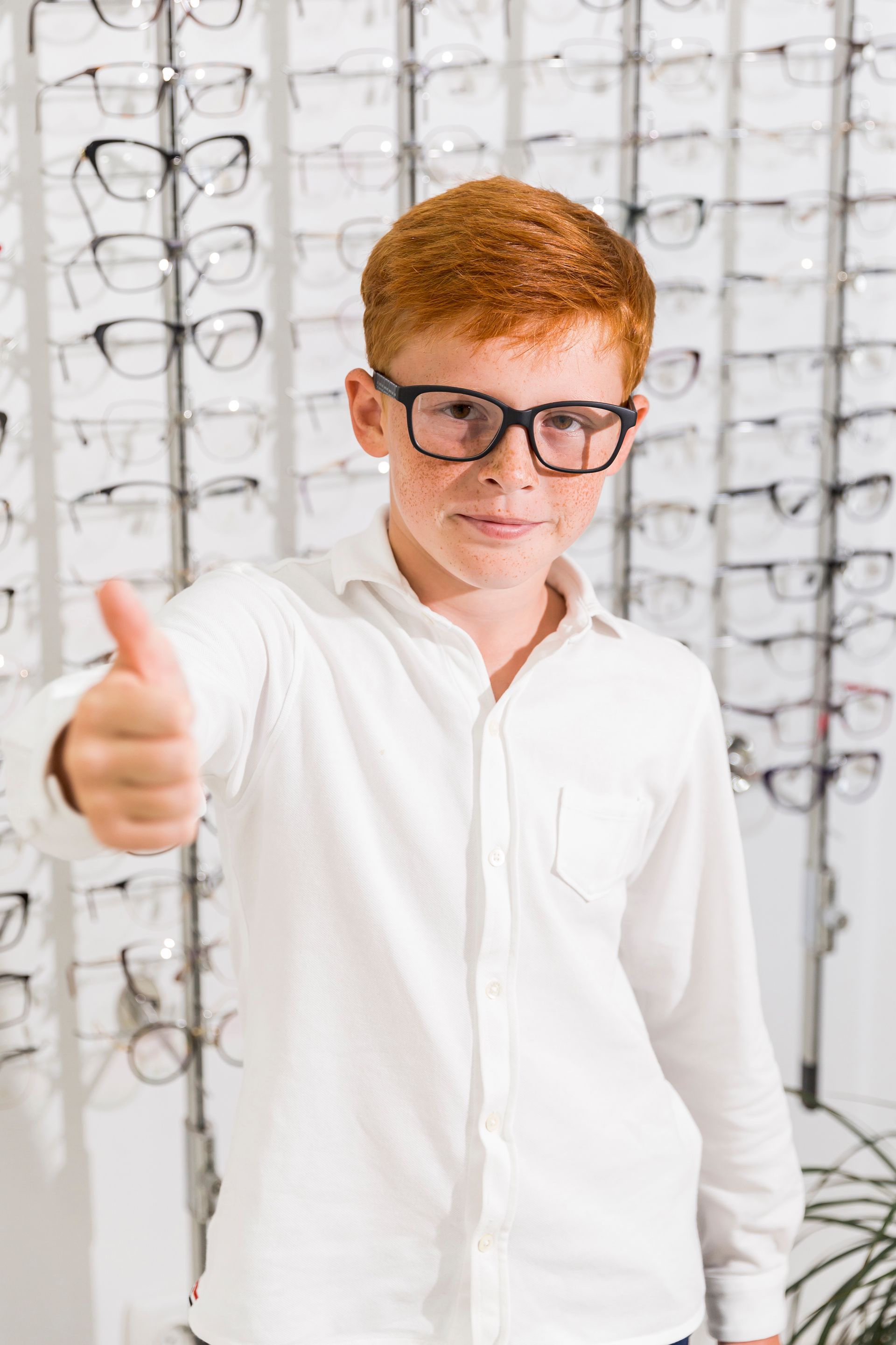 A young boy wearing glasses is giving a thumbs up in an optical shop.