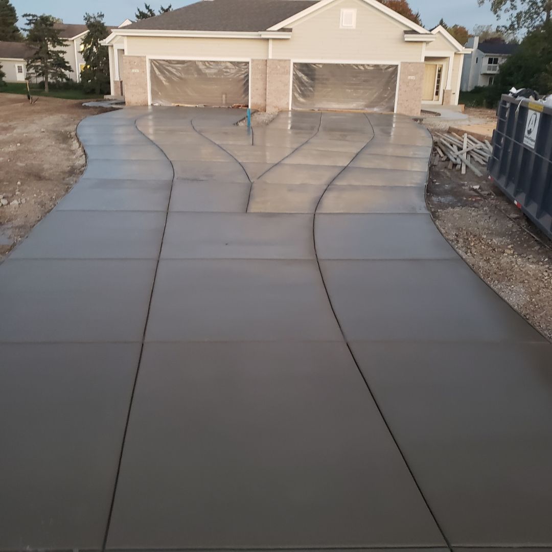 Freshly poured concrete driveway leading to a beige house with two garage doors, still edged by gravel.