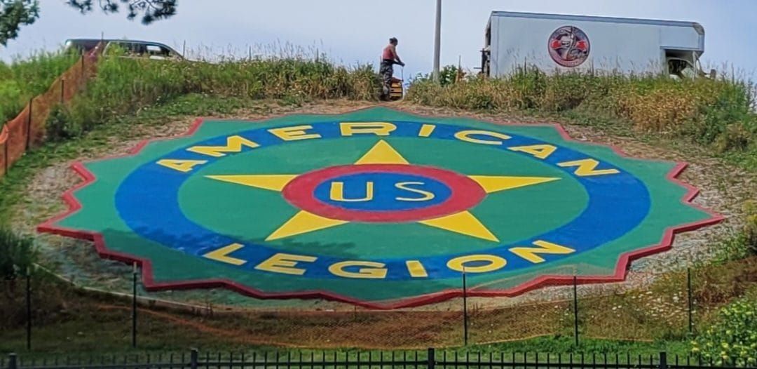 Large colorful circular seal painted on ground reading “Marines Legion,” with a truck and people in the background