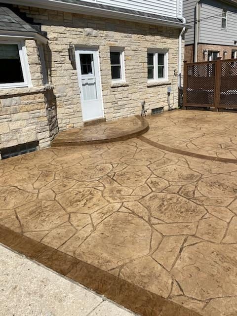 Stamped concrete patio and steps beside a stone house with a white door and wooden fence.