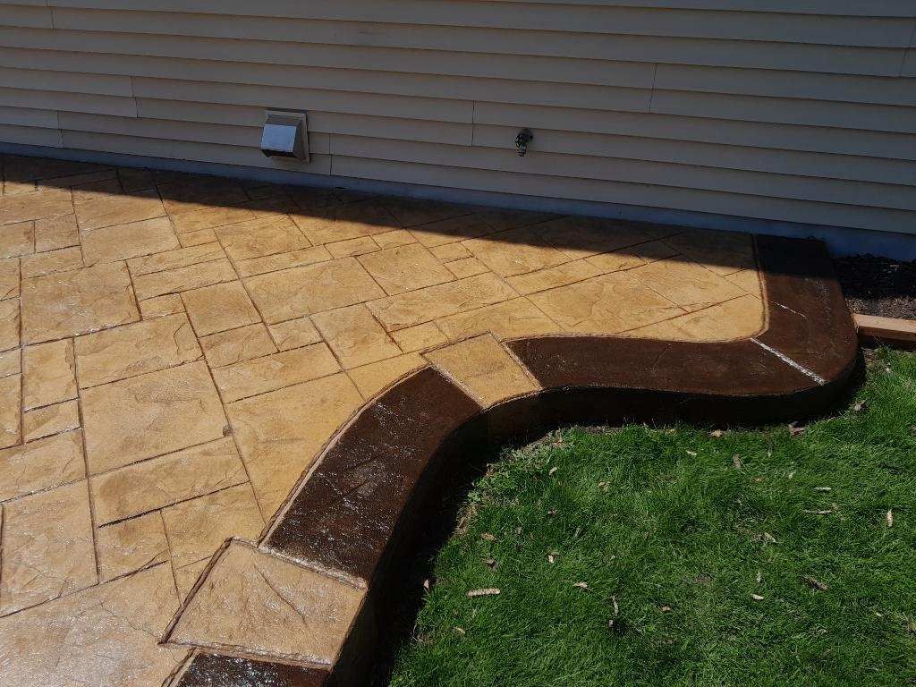 Stamped concrete patio with a curved dark border beside green grass and house siding.